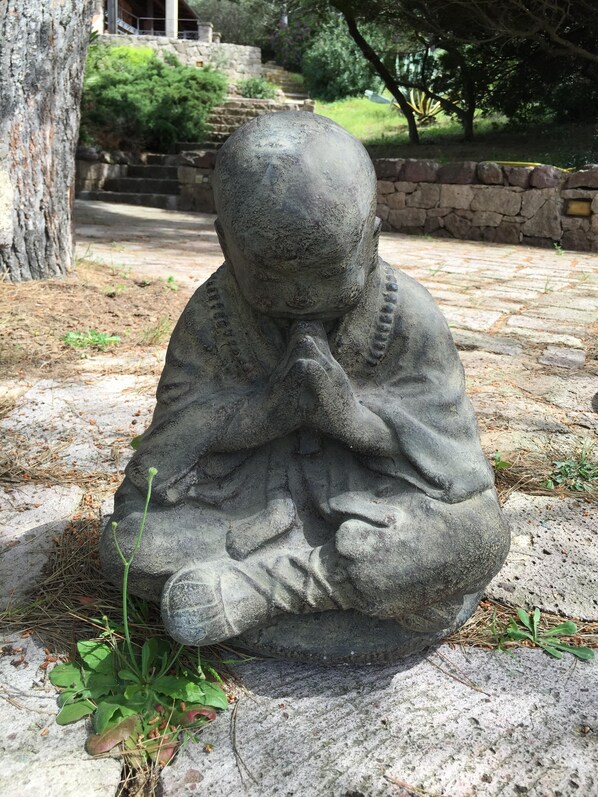 A stone statue of a seated figure with hands clasped in prayer, surrounded by greenery and a stone wall in the background.