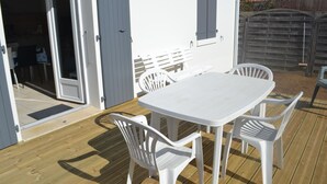 Une terrasse extérieure avec une table et des chaises blanches, une plateforme en bois et un bâtiment blanc doté d’une porte et d’une fenêtre à volets.