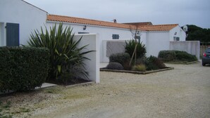 Un bâtiment blanc avec un toit recouvert de tuiles, une allée gravillonnée et diverses plantes.