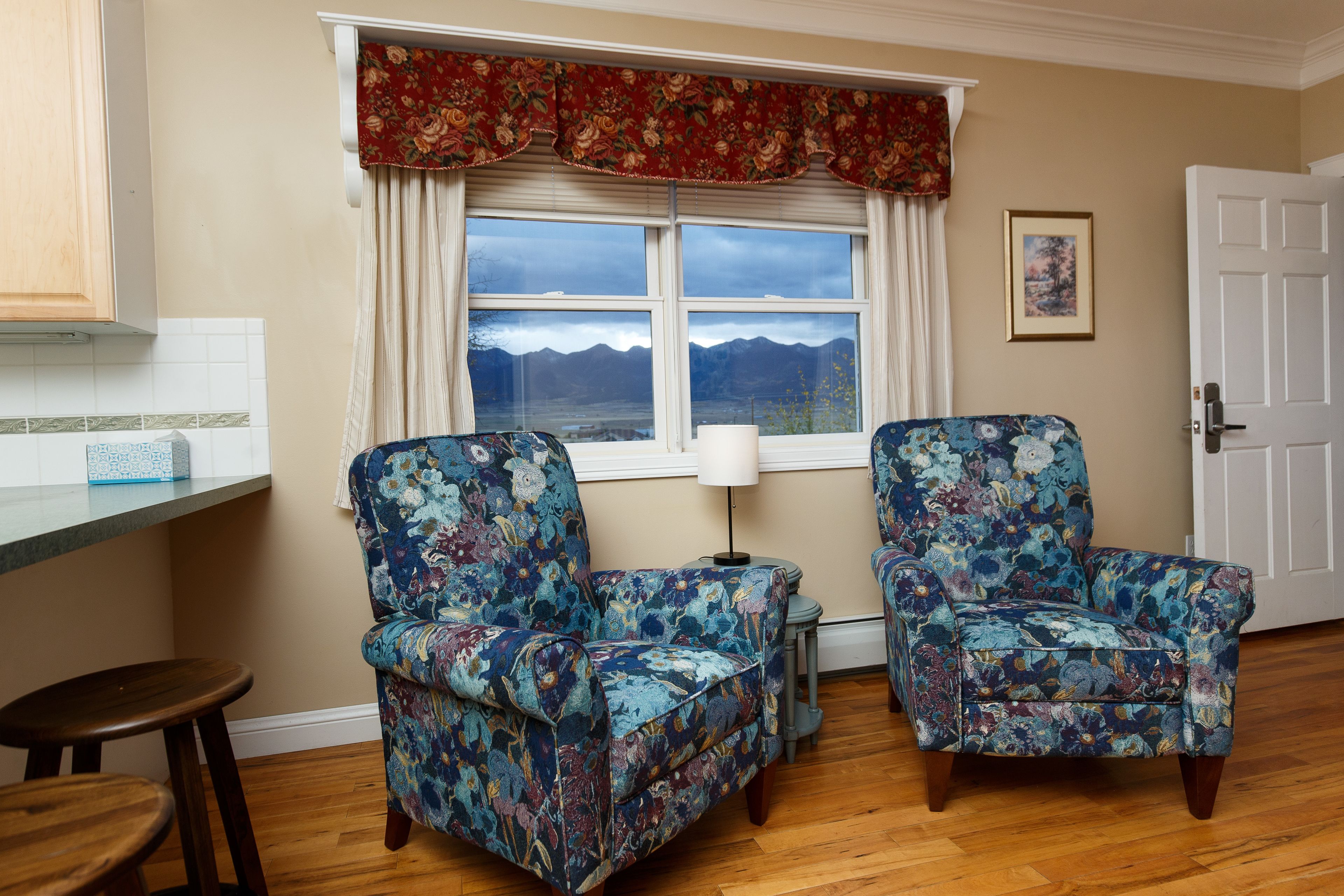 Ein Zimmer mit zwei floralen Sesseln, Holzboden, einem Fenster mit Bergblick, einer Tür und einem kleinen Tisch mit einer Lampe.