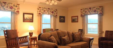 A living room with a brown sofa, a wooden rocking chair, and a patterned armchair. There are two windows with floral curtains, a ceiling fan, and a lamp on a side table.