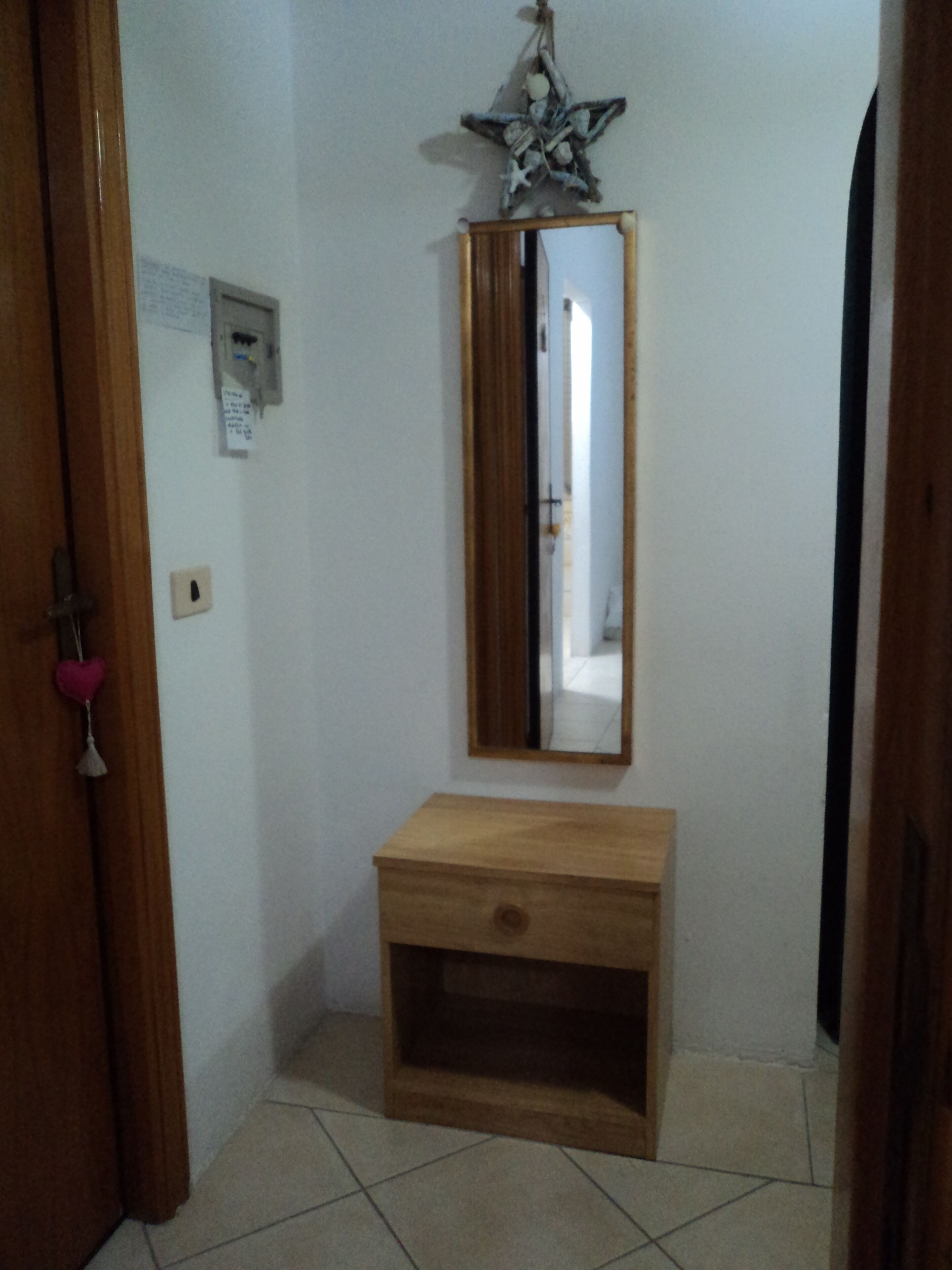 Une coiffeuse en bois avec un miroir au-dessus, une petite étagère et un luminaire fixé au mur.