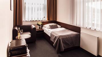 Habitación de hotel compacta con una cama individual, un pequeño sofá, una mesita de noche con lámpara, un jarrón con flores y una maleta sobre un carrito.