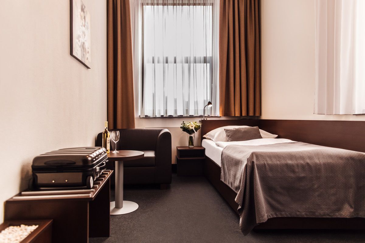 A hotel room with a bed, a sofa, a small table with a lamp, a suitcase, and a vase with flowers.