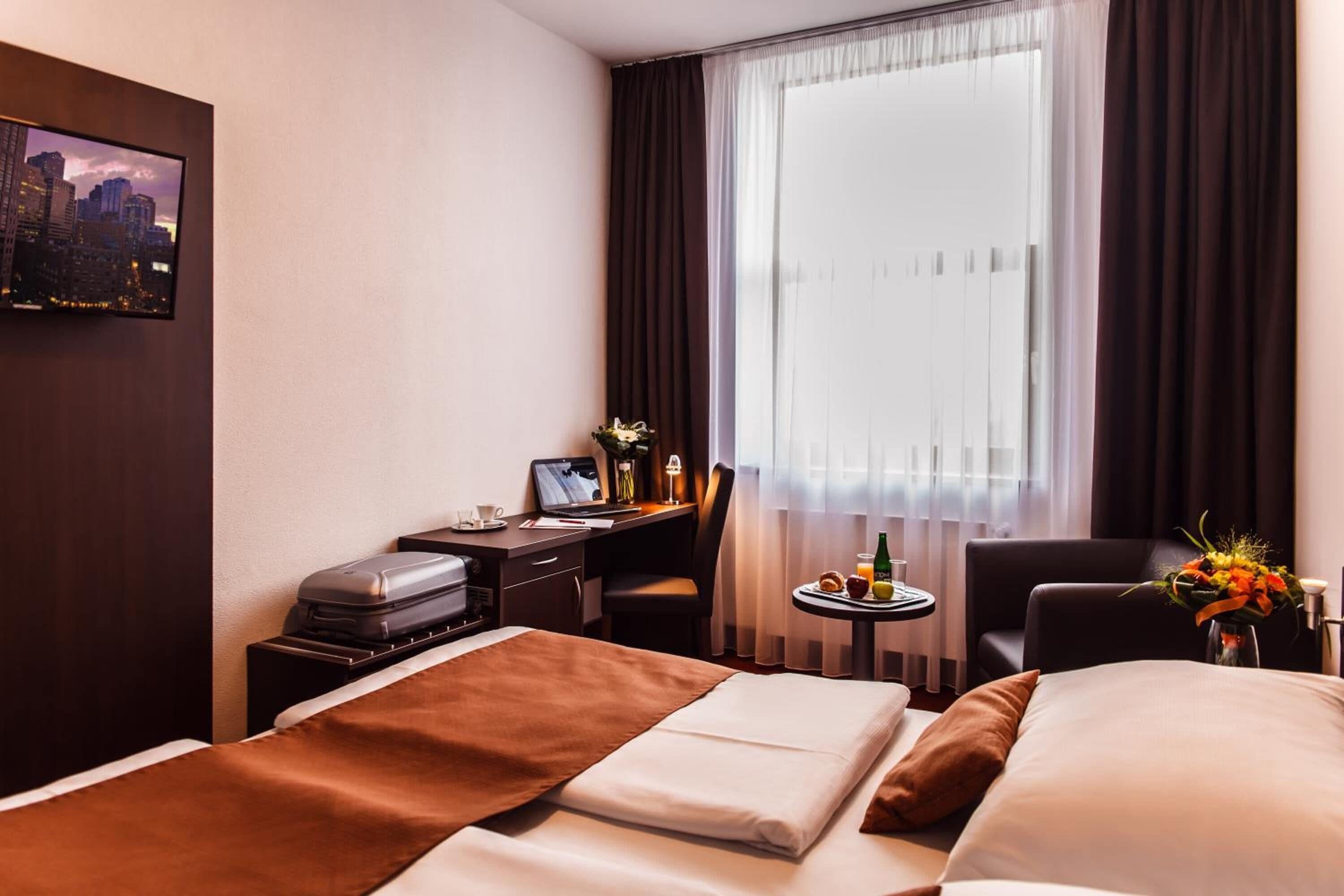 Ett hotellrum med en säng, ett skrivbord med en bärbar dator, en stol, ett litet bord med ett glas och ett fönster med gardiner.