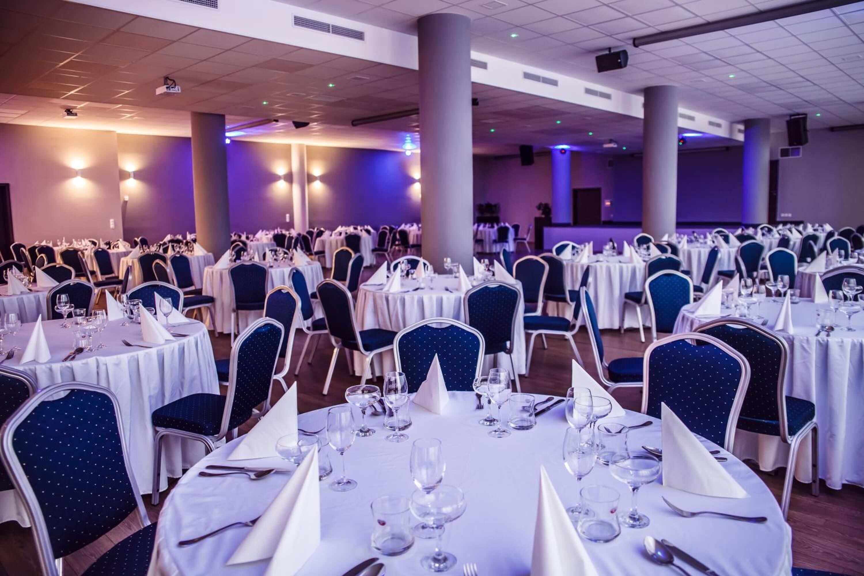 Un salón de banquetes con mesas redondas dispuestas para cenar, cada una con mantel blanco y sillas azules.