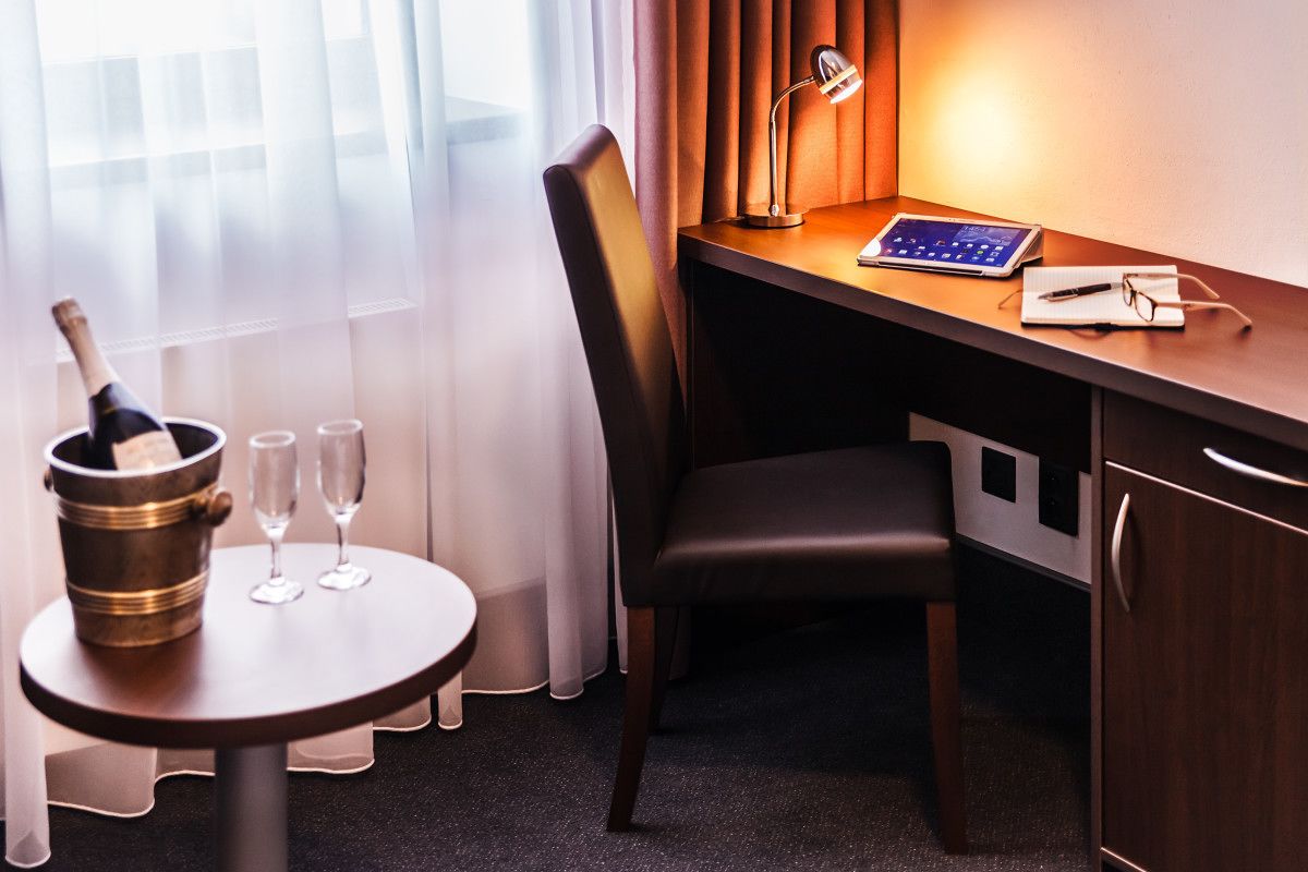 A hotel room with a desk, chair, and a small table with a bottle of champagne and two glasses.