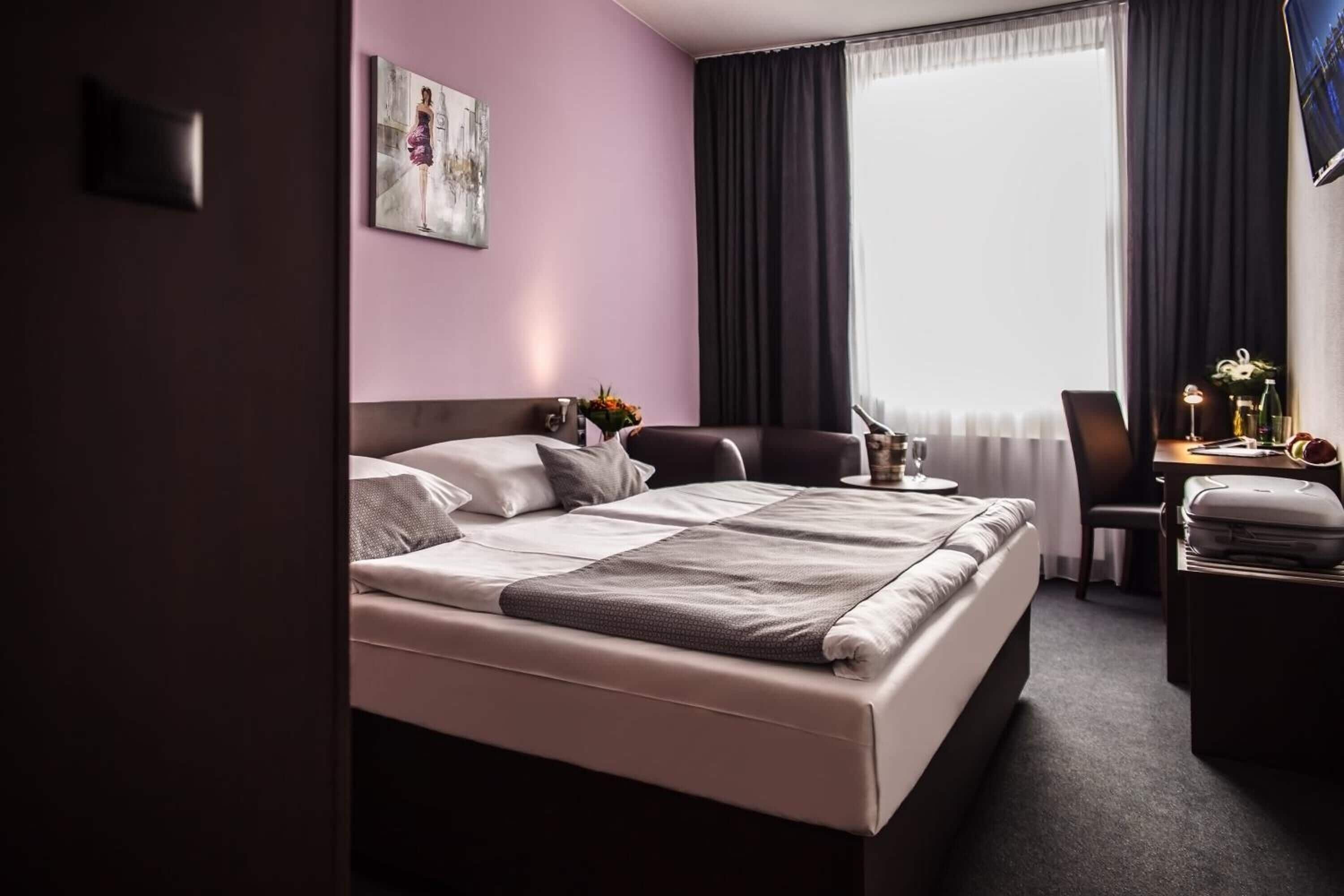 A modern hotel room with a large bed, a desk, and a chair. There is a window with curtains, a painting on the wall, and a vase with flowers on the desk.
