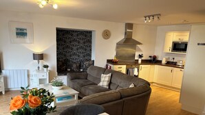 A modern living room with a brown sofa, a dining table with a vase of flowers, and a kitchen area with white cabinets and a stainless steel range hood. - Magnificent Sea views, Central, 2 double bedrooms, 2 bathroom. Pets welcome.   (Newquay)