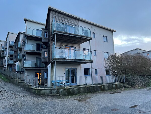Un edificio residencial de varios pisos con balcones, una entrada protegida por un portón y un camino de entrada de grava.