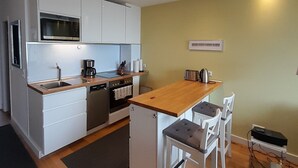 Cocina compacta con armarios blancos, horno empotrado y microondas. En la encimera, hay un refrigerador pequeño, una cafetera y un hervidor. El piso es de madera y cuenta con una pequeña zona de comedor con dos sillas.