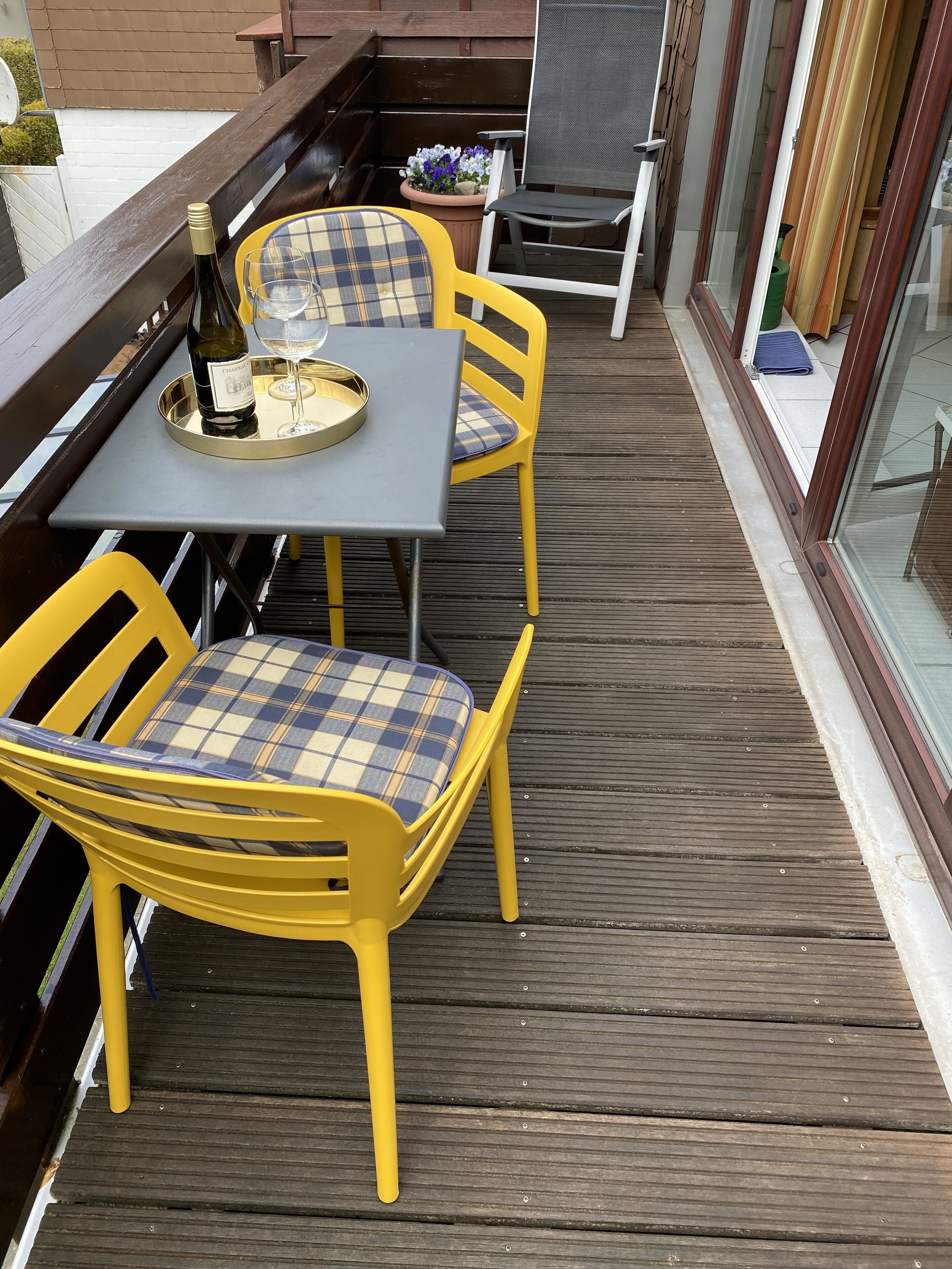 Une terrasse en bois avec des chaises jaunes et une table, une bouteille de vin et un verre posés sur la table.
