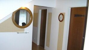 A hallway with a wooden floor, a round mirror on the left, a clock on the right, and a door at the end.