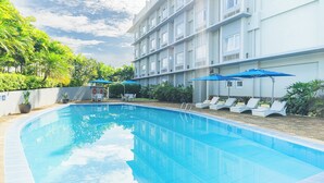 Una piscina con sillones de descanso y sombrillas, rodeada de follaje y un edificio de varios pisos.