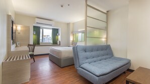 Una habitación de hotel moderna con sofá, cama, suelo de madera, ventana con cortinas y aire acondicionado de pared.