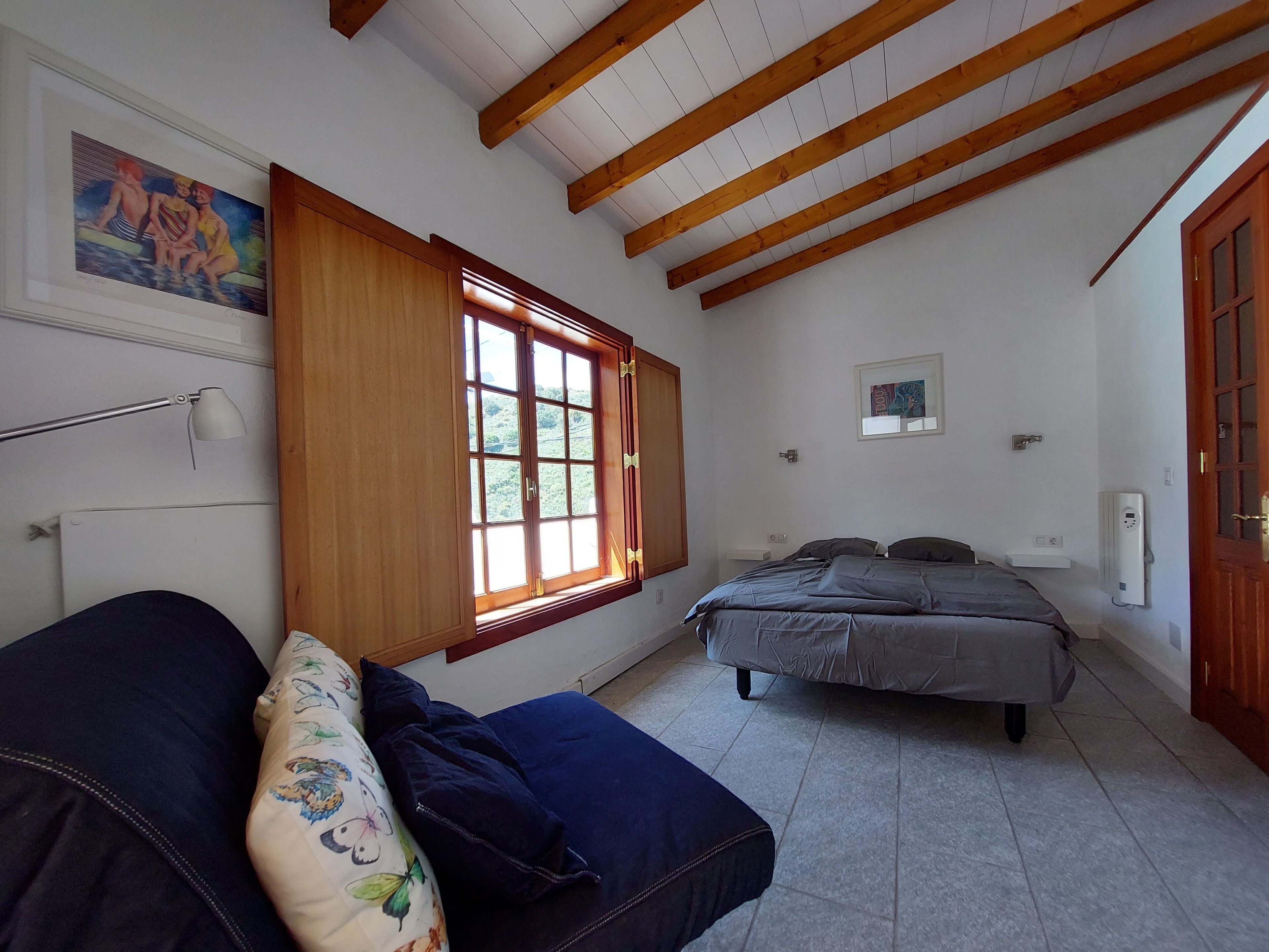 Een slaapkamer met een bed, houten plafond, een raam, een wandlamp en een schilderij.