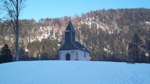 Una piccola chiesa in pietra con un campanile a guglia, immersa in un paesaggio innevato e con una collina boscosa sullo sfondo.