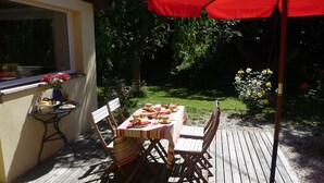 Un espace de restauration en plein air avec une table dressée pour deux personnes, entourée de chaises en bois et d’un parasol rouge.