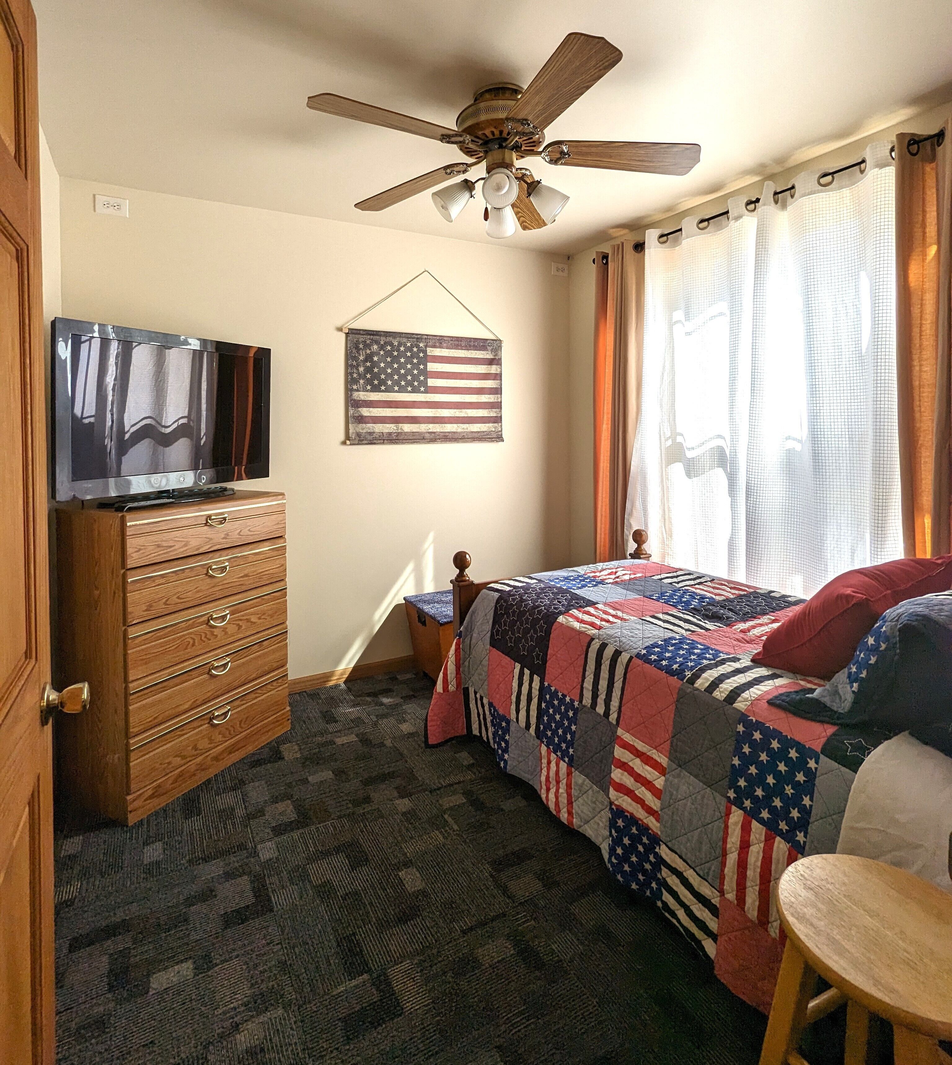 A bedroom with a bed, a dresser, a ceiling fan, a TV, and an American flag.