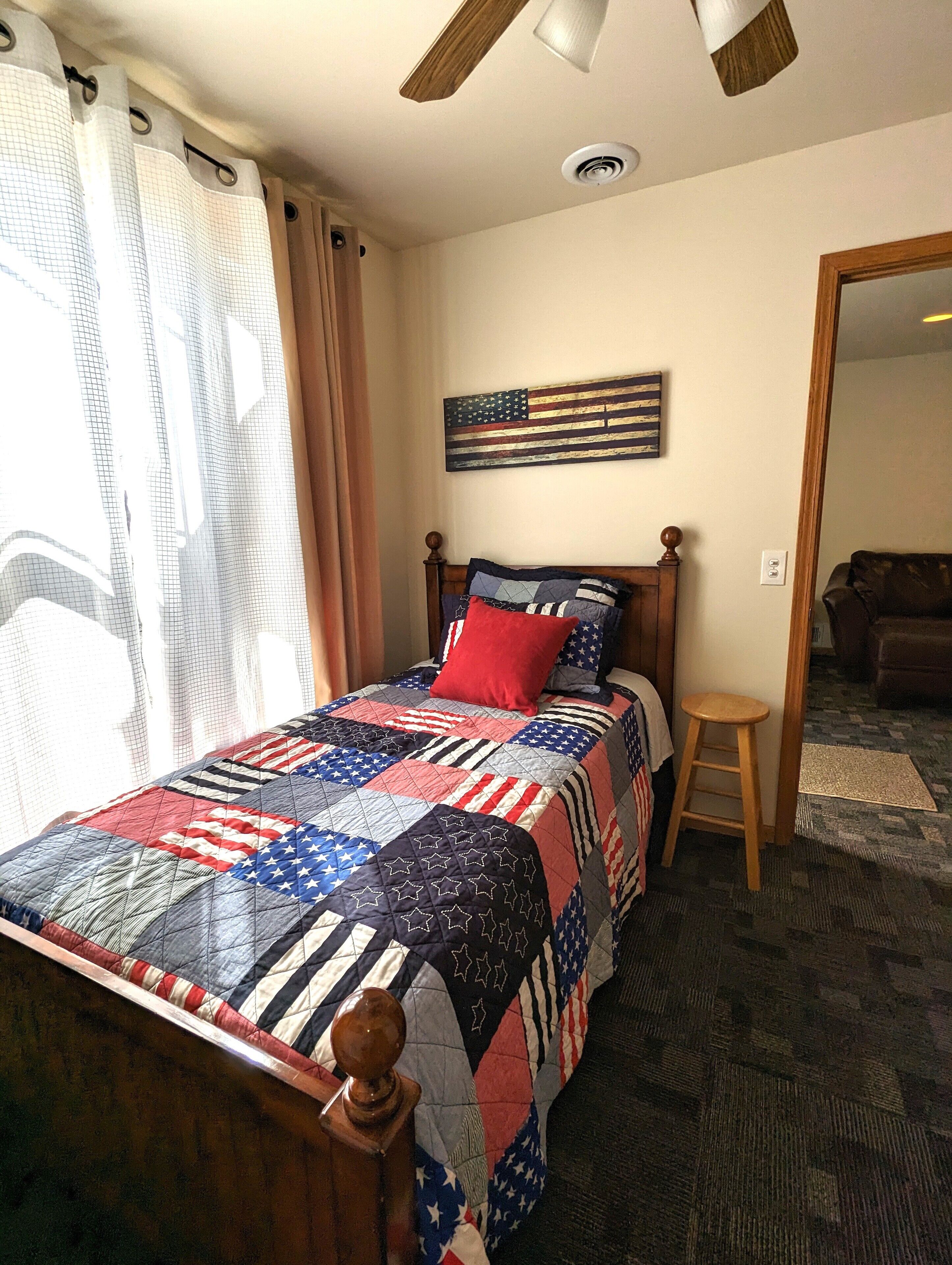 Un dormitorio con cama, cabecera de madera, almohada roja, colcha con estampado de estrellas, ventilador de techo y puerta que da a otra habitación.