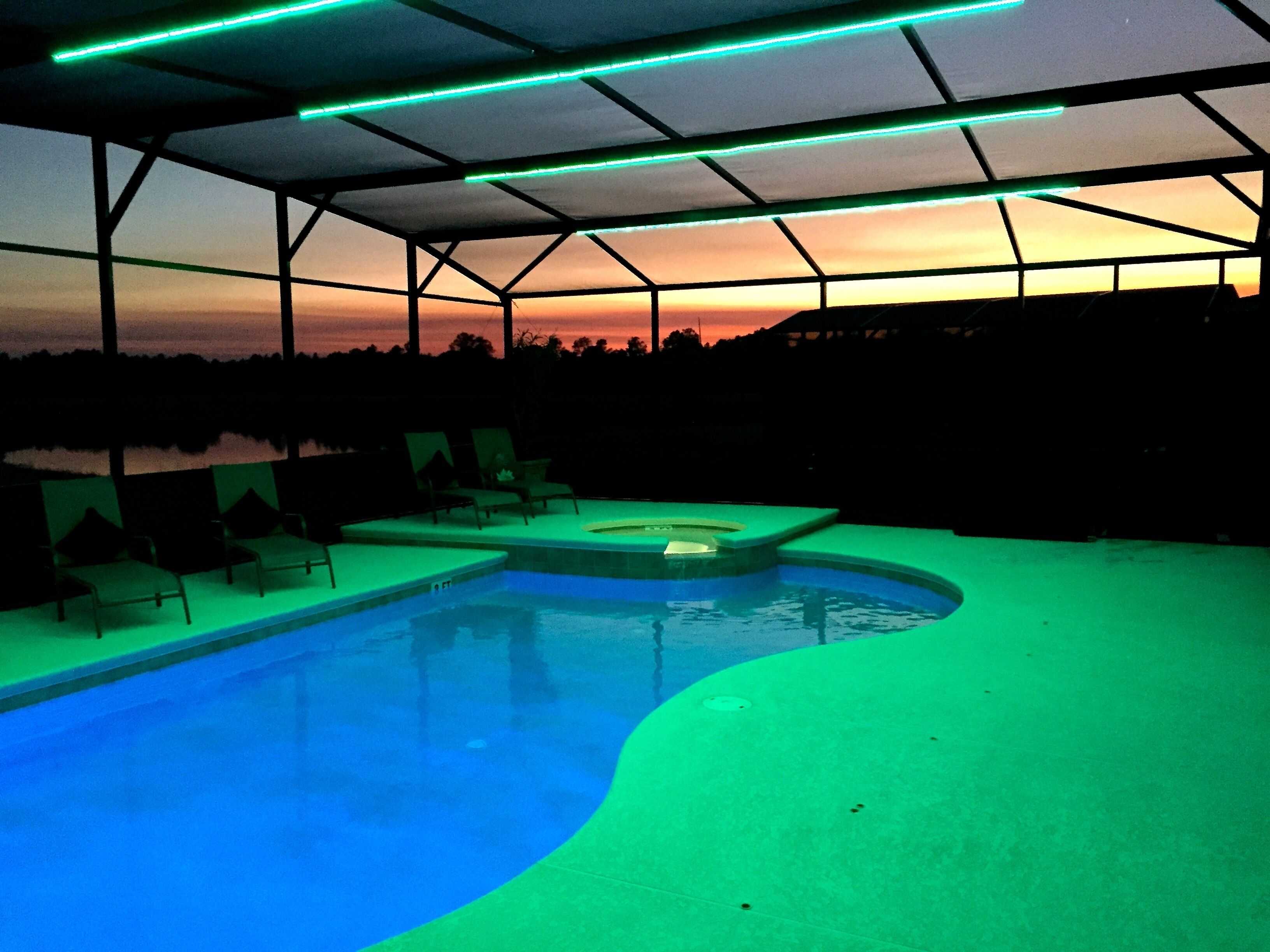 A swimming pool with a hot tub, surrounded by lounge chairs, under a roof with green lighting.