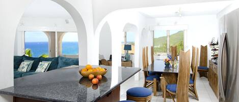 A modern kitchen with a central island, a dining area with wooden chairs, and a view of the sea.