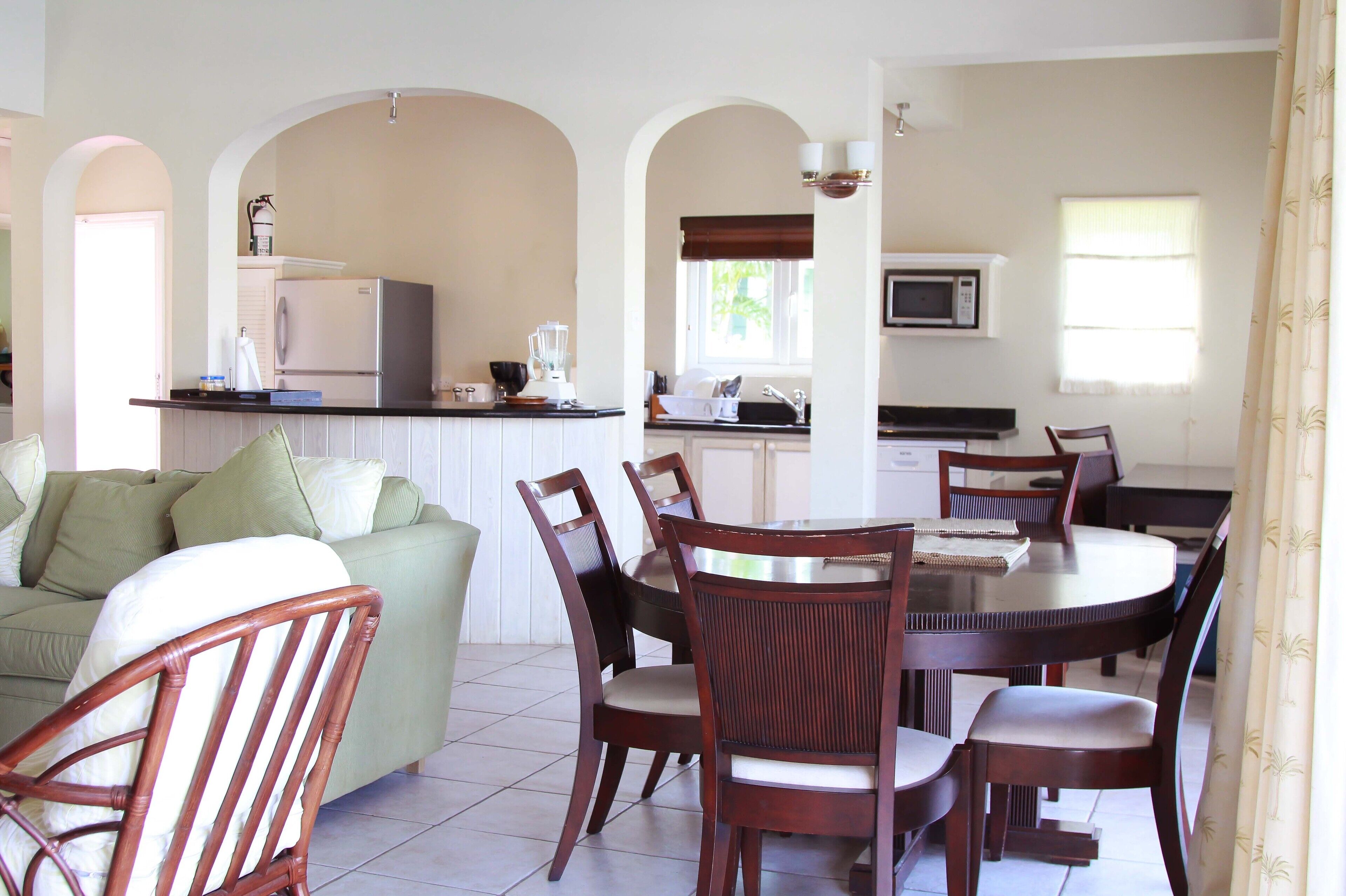 A dining area with a round table and chairs, a kitchen with a refrigerator and microwave, and a living area with a sofa and chairs.