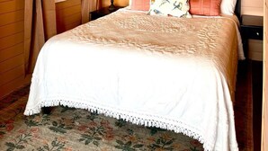 A cozy bedroom with a wooden ceiling, a bed with floral bedding, a round mirror, and a window with curtains. - Snug Harbor Inn - Midship Cottage (Sturgeon Bay)