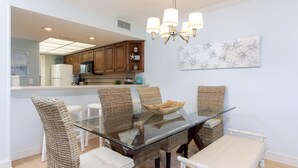Un coin repas avec une table à plateau en verre entourée de chaises en osier, un lustre et une cuisine en arrière-plan.