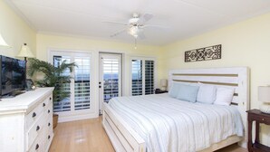 Une chambre à coucher avec un lit, un ventilateur de plafond, un téléviseur posé sur une commode et une plante.