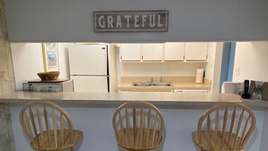 Een keuken met een witte koelkast, een spoelbak en houten barstoelen. Een bord boven de toonbank met de tekst 'GRATEFUL'.