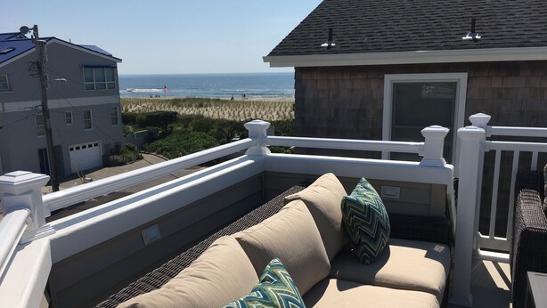 A balcony with a beige sofa and cushions, overlooking a beach and ocean view.
