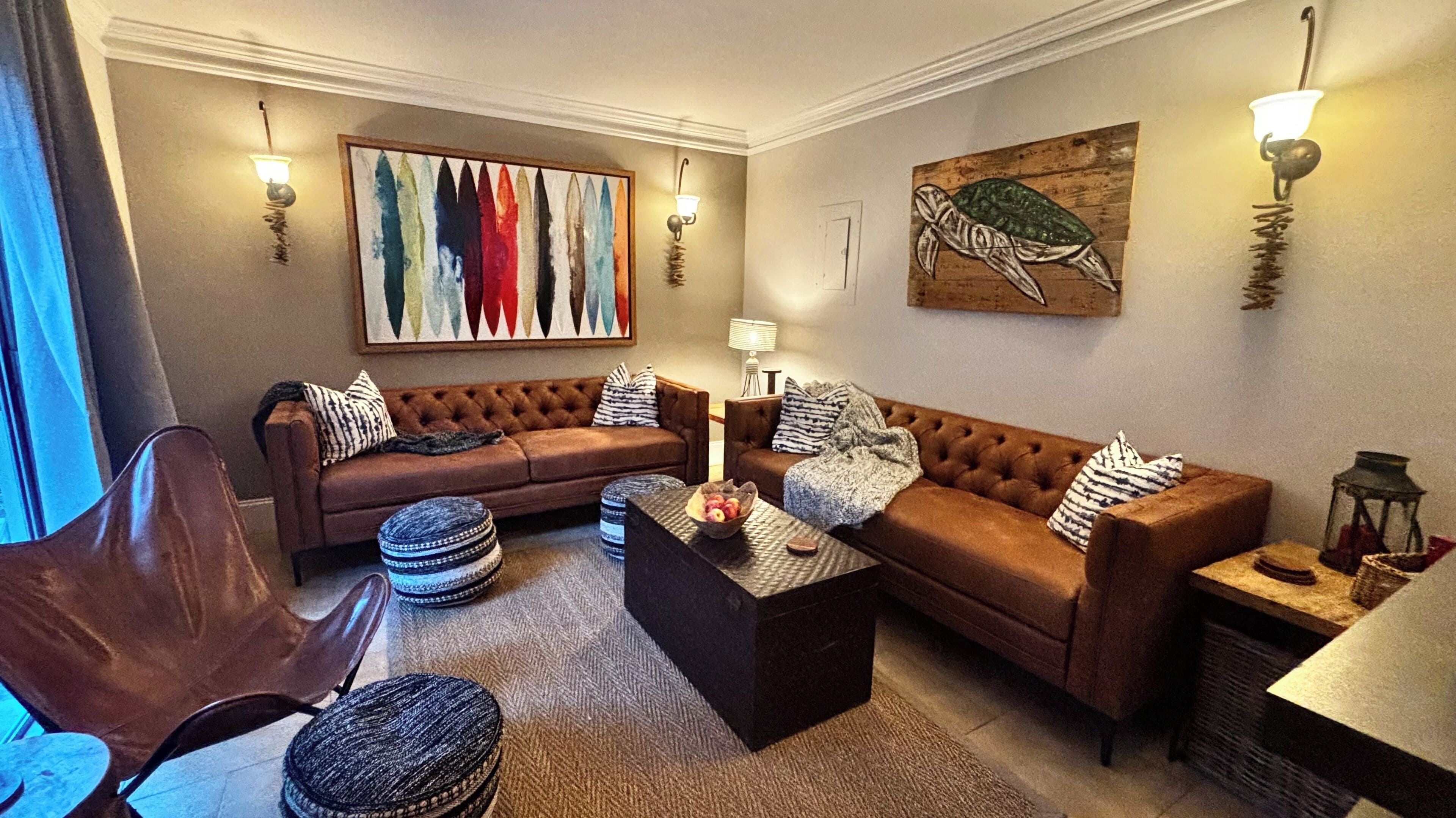 Een woonkamer met een bruine bank, een salontafel en een fauteuil. Er zijn wandlampen en een schilderij aan de muur.