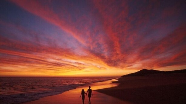 Un atardecer sobre una playa con dos siluetas caminando.