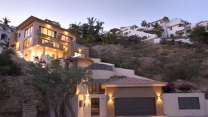 Una casa moderna de dos pisos con garaje, rodeada por un paisaje montañoso y palmeras.