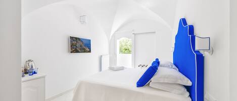 A modern bedroom with a white and blue color scheme, featuring a bed, a bedside table, a window, and a view of the outdoors.