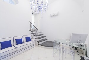 Un salón moderno con un sofá blanco, cojines azules, un escritorio de cristal y una escalera.