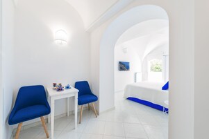 Una habitación moderna con una mesa blanca, dos sillas azules, una cama con ropa de cama azul y un amplio arco de entrada a otra habitación.