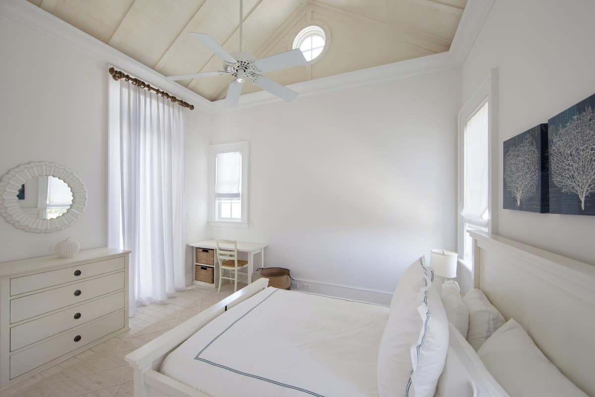 A bedroom with a bed, a desk, a chair, a ceiling fan, and a window.