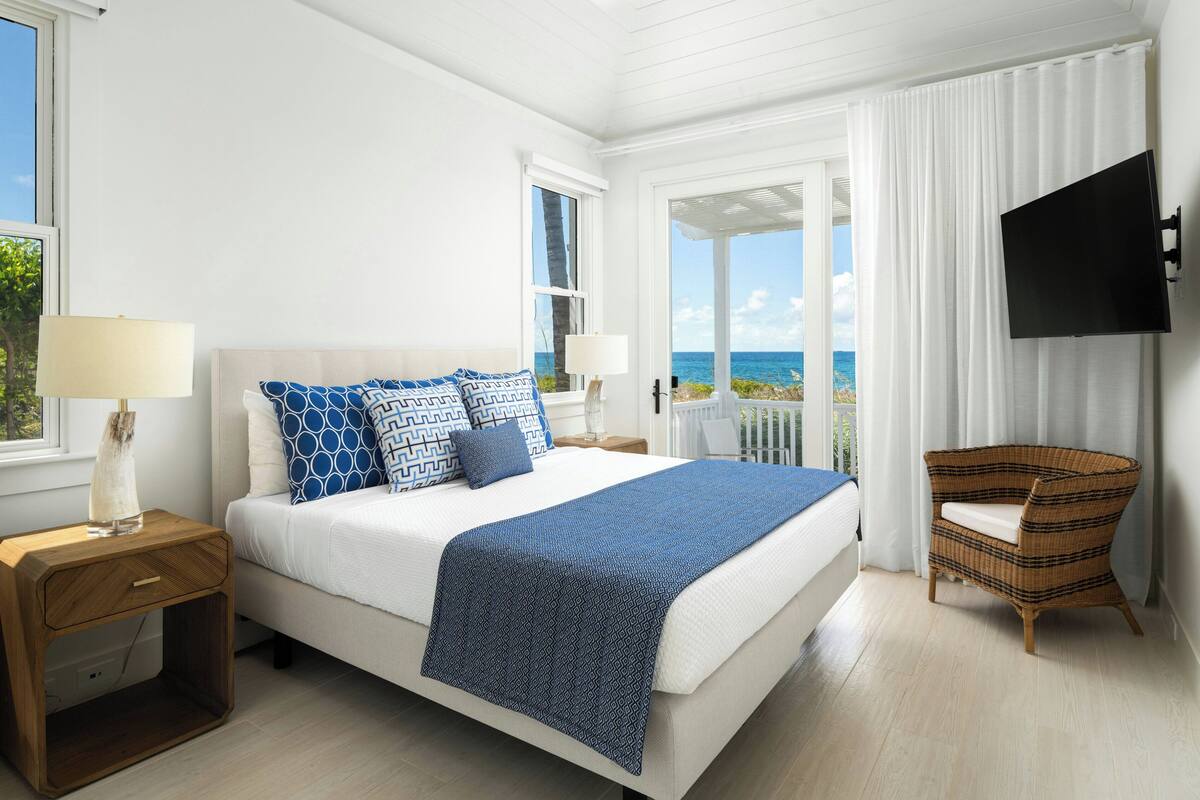 A bedroom with a bed, two nightstands, a wicker chair, a TV, a window with curtains, and a view of the ocean.