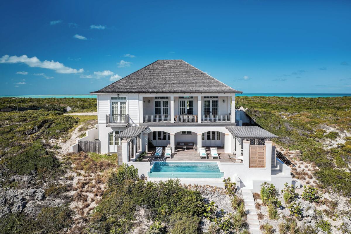A large white house with a swimming pool, multiple windows, and a balcony overlooking a beach.