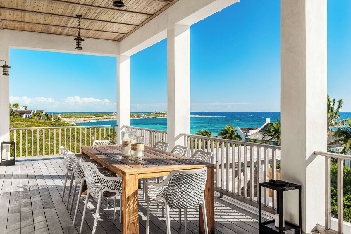 A covered outdoor area with a wooden table and chairs, a white railing, and a view of the ocean.