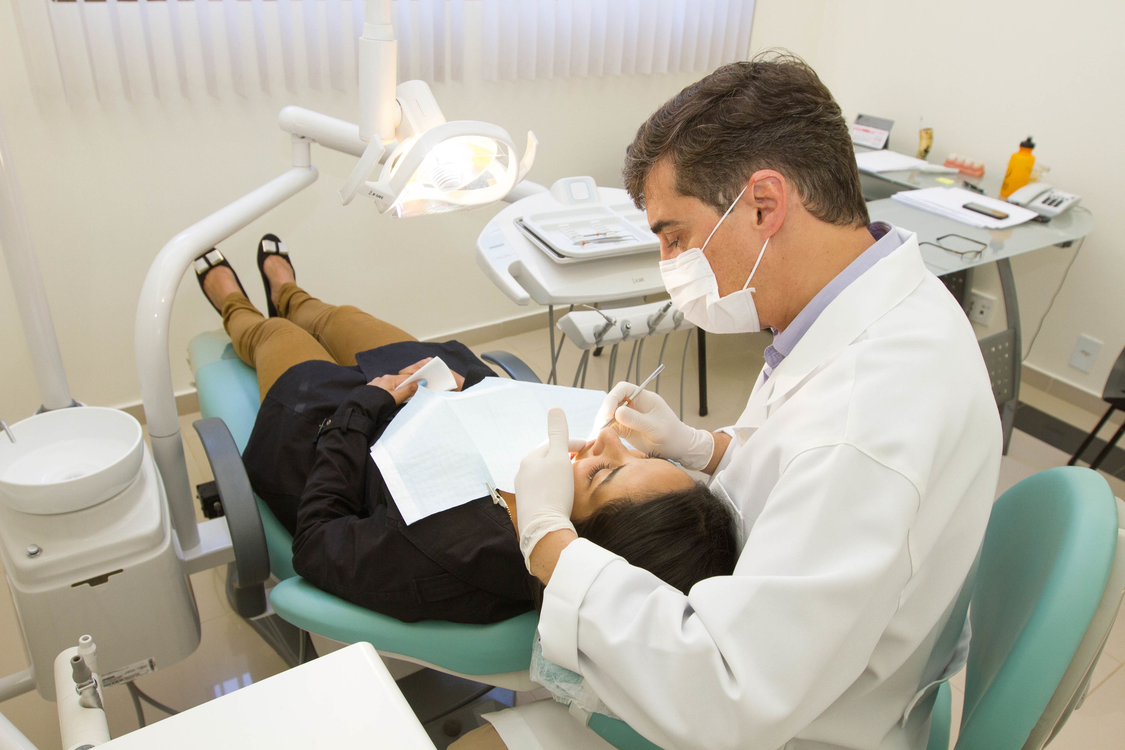 Clínica dentária com um paciente deitado numa cadeira de dentista, um dentista usando máscara e equipamentos dentários sobre uma mesa.