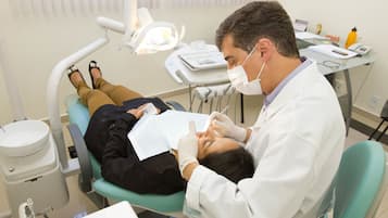 Clínica dentária com um paciente deitado numa cadeira de dentista, um dentista usando máscara e equipamentos dentários sobre uma mesa.