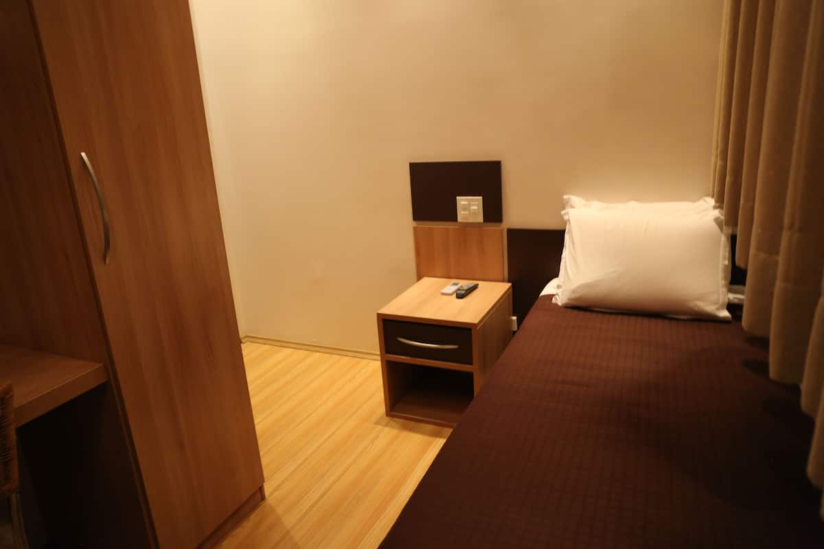 Une chambre d’hôtel avec une armoire en bois, une petite table de chevet en bois avec une télécommande, un lit avec un oreiller blanc et un plancher en bois.
