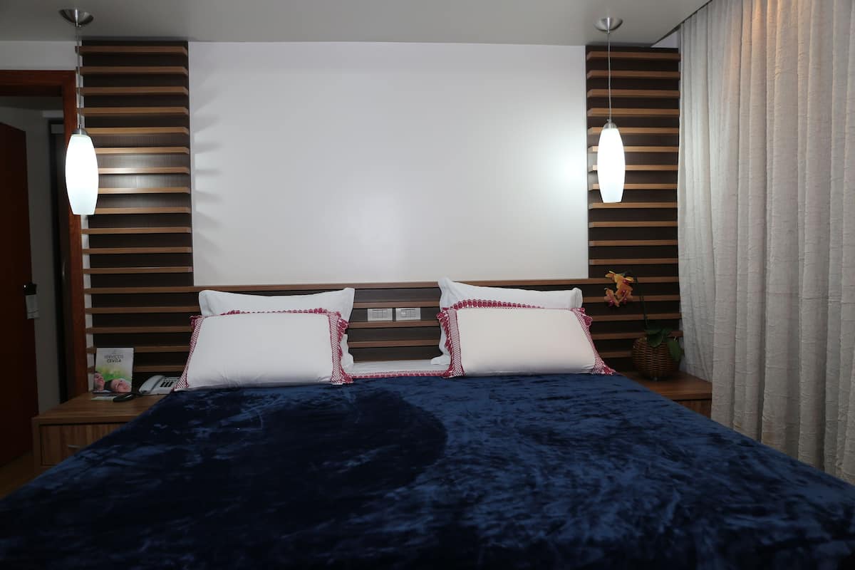 Un lit bien fait, recouvert d’une couverture bleu foncé, avec deux oreillers blancs à bordure rouge et une tête de lit en bois. Deux lampes suspendues sont accrochées au-dessus du lit, et une petite table, sur laquelle se trouvent une plante et un téléphone, est placée à côté.