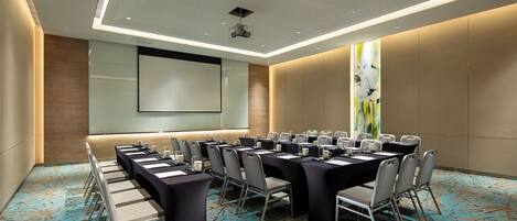 A conference room with long tables, chairs, a projector screen, and a large abstract painting on the wall.