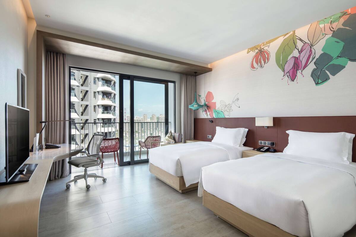 A hotel room with two beds, a desk with a TV, a chair, and a large window with a view of a building.