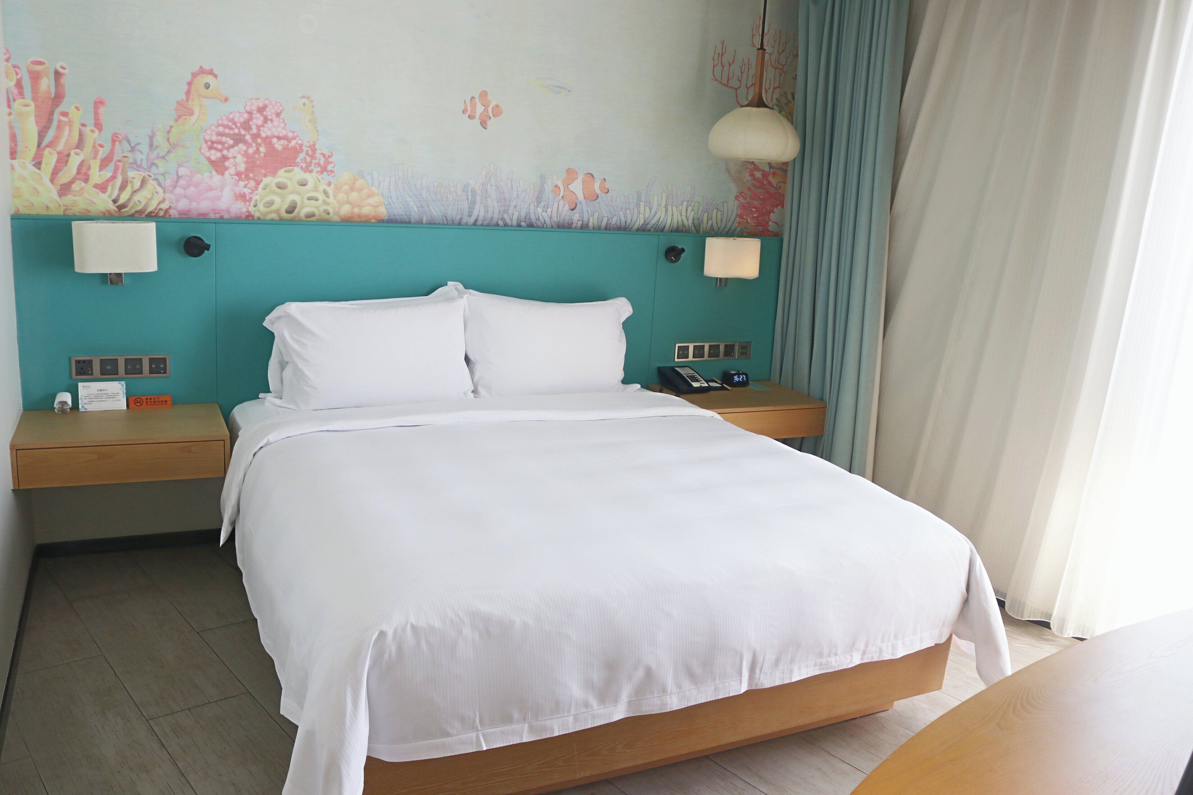 Una habitación de hotel con cama, mesitas de noche, un mural en la pared y una ventana con cortinas.
