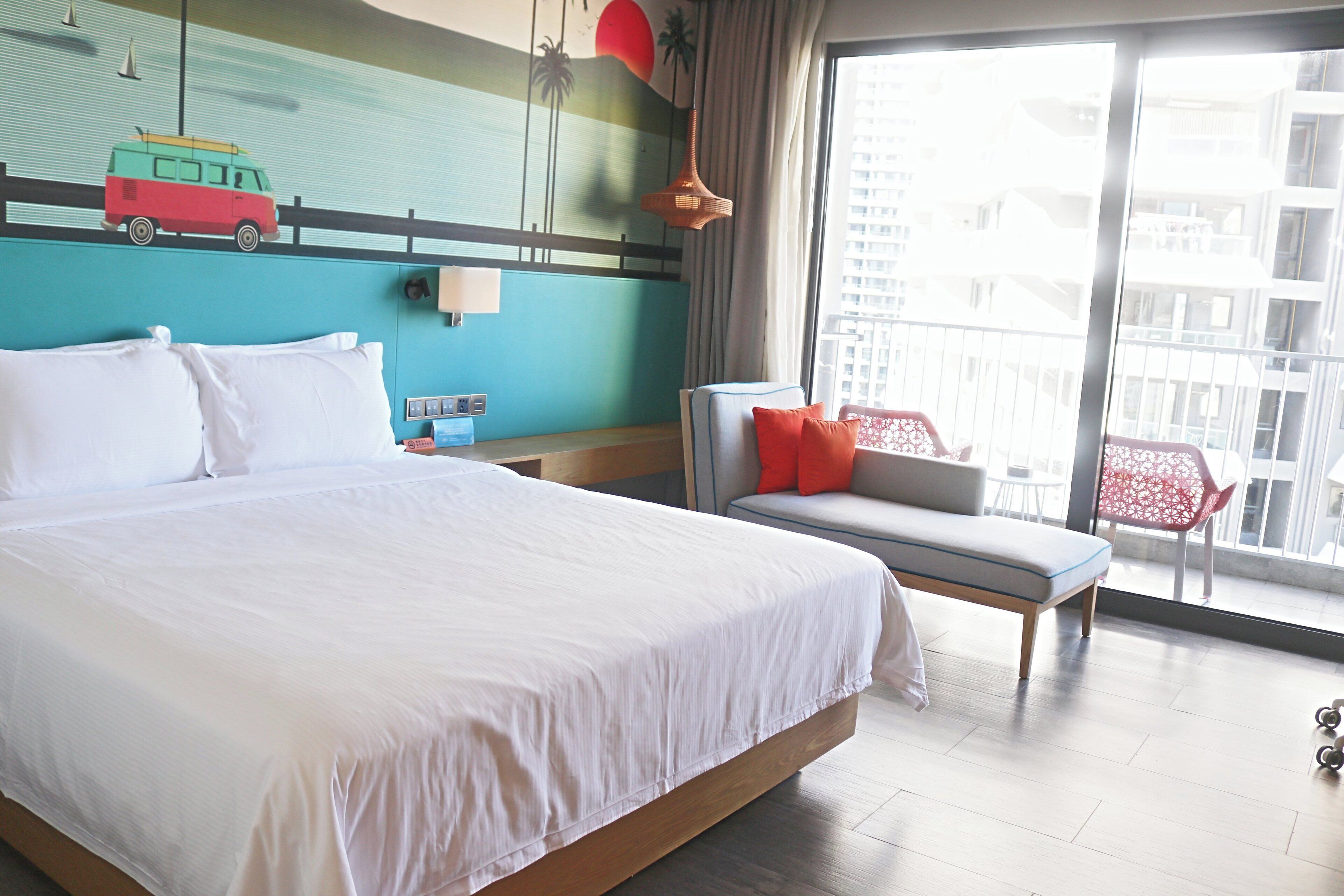Ein Hotelzimmer mit einem großen Bett, einer kleinen Couch, einem Fenster mit Blick auf Gebäude und einer Wand mit Wandgemälde.